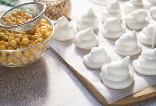 Vegane Baisers auf einem Holzbrett, daneben Kichererbsen und Aquafaba in einer Schüssel