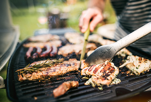 Helles Fleisch auf einem Grill wird von einem Mann gewendet, im Hintergrund ist ein Garten.