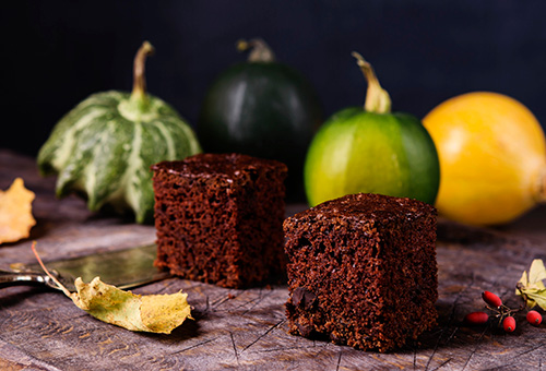 Zwei Stücke Zucchini-Brownie auf einem Tisch, im Vorder- und Hintergrund Tischdekoration aus Zierkürbissen und Laub