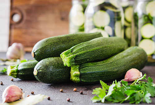 wissenswert-grüene-zucchini-einkochen Grüne Zucchini mit Knoblauchzehen, Kräutern und Pfeffer auf einer dunklen Arbeitsplatte mit gefüllten Einmachgläsern im Hintergrund