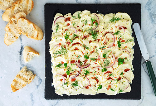 angerichteter Dattel Cranberry Dip garniert mit roten Zwiebeln auf einer Schieferplatte mit Baguettescheiben und einem Messer 