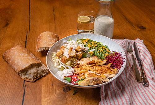 Ceasars Salad Bowl mit Avocado in Teller angerichtet plus Ciabatta und Dressing separat daneben auf Holztisch