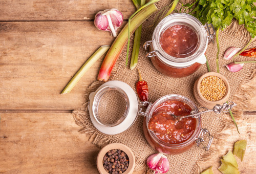 Handelshof Rezept Rhabarberchutney Rhabarberchutney in Gläsern und Knoblauchzehe und Pfefferkörner in Schüssel und Chilis auf einer Tischplatte