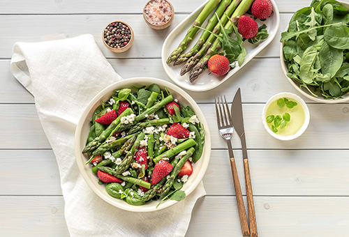 Warmer Spargel mit Erdbeeren, Rucola und Vinaigrette angerichtet in einer weißen Schüssel auf einem Tisch mit Besteck und einer Schale mit Salat sowie einer Platte mit grünem Spargel