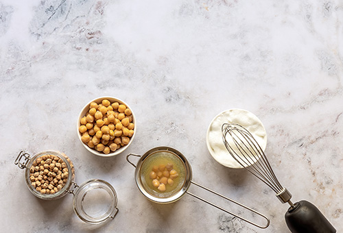 Von der Kichererbse zu veganem Eischnee mit Aquafaba Getrocknete Kichererbsen, gekochte Kichererbsen, Aquafaba und veganer Eischnee in Glasschüsseln