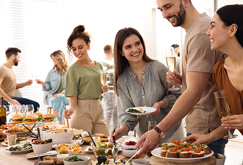 Menschen stehen an einem Brunchbuffet und nehmen sich Essen und halten Sektgläser in den Händen