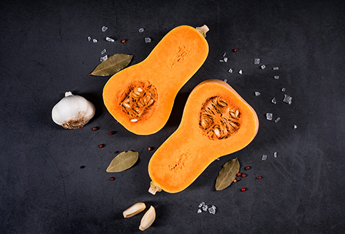 Halbierter Butternutkürbis liegt auf einem schwarzen Hintergrund, drum herum liegen große Salzflocken und eine Knoblauchzehe als Dekoration