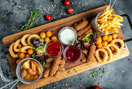 Verschiedene frittierte Speisen, wie Pommes und Zwiebelringe und Mozzarellasticks, auf einem Holzbrett mit Dips