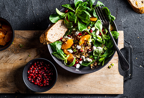 Salat mit Granatapfelkernen Feldsalat mit Orangen und Granatapfelkernen in einer schwarzen Schüssel, daneben eine Schüssel mit Granatapfelkernen, alles steht auf einem Holzbrett