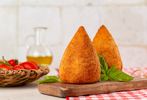 Zwei Arancini auf einem Holzbrett, im Hintergrund Tomaten in einem Korb und eine Flasche Olivenöl