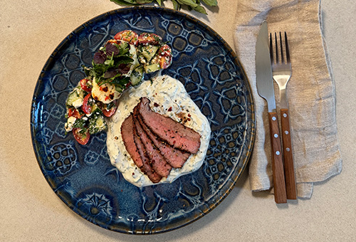 Gegrilltes Flanksteak mit Hirtensalat sowie gegrillter Halloumi Gegrilltes Flanksteak mit Hirtensalat sowie gegrillter Halloumi mit Petersilien-Minz-Salat und Tsatsiki auf einem blauen Teller angerichtet