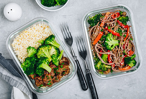 Zwei Glasschüsseln mit vorbereitetem Essen. Einmal Soba-Nudeln mit Brokkoli und Paprika und einmal Brokkoli, Reis und Hühnchen.