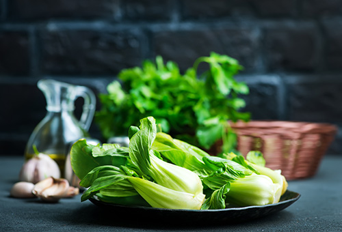 Pak Choi in der Küche Zwei Pak Choi auf einem schwarzen Teller, im Hintergrund Olivenöl und Knoblauchzehen als Dekoration