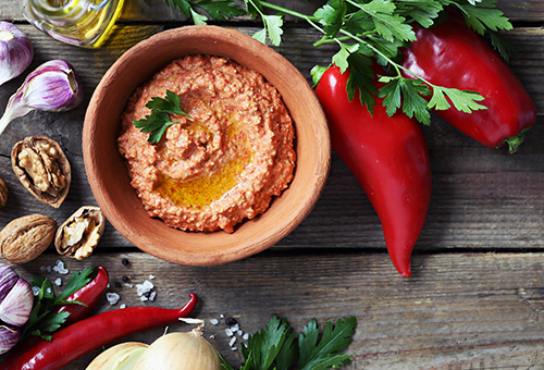 Muhammara Muhamarra in einer Schale angerichtet auf einem Tisch, der mit Peperoni, Walnüssen und Knoblauch dekoriert ist