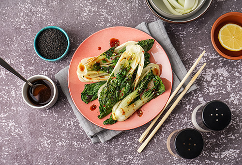 Pak Choi mit würziger Soße Pak Choi mit würziger Soße beträufelt auf einem pfirsichfarbenen Teller, rundeherum Salz, Pfeffer, Öl und schwarzer Sesam als Dekoration
