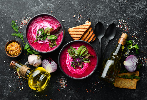 Cremige Rote Bete Suppe in zwei schwarzen Suppentellern auf einem dunklen Tisch, der mit geröstetem Brot, Löffeln und Olivenöl dekoriert ist
