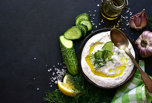 Tsatsiki in einer Schüssel, daneben Gurke, Knoblauch, Zitronenspalten, Dill, Salz und eine Karaffe mit Olivenöl als Dekoration
