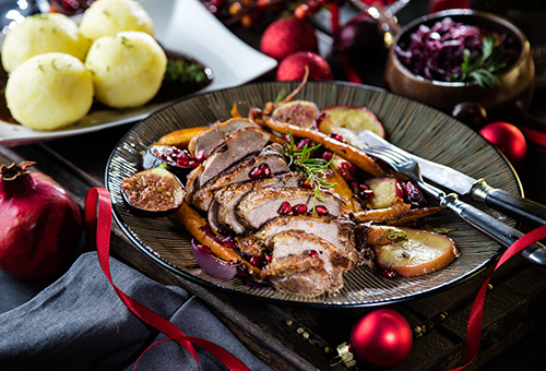 Entenbrust mit Birne und Granatapfel auf einem grauen Teller angerichtet. Teller steht auf einem festlich dekorierten Tisch, im Hintergrund sind Klöße zu sehen