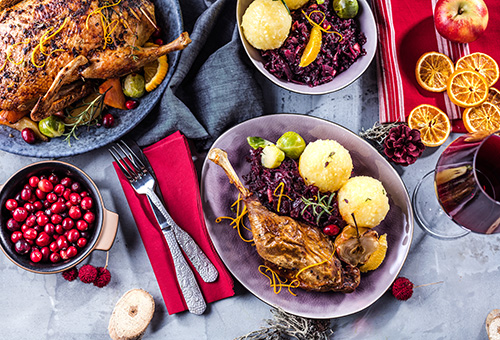 Gänsekeule in einer grauen Schüssel mit Klößen und Rotkraut auf einem festlich dekorierten Tisch, im Hintergrund ist die Gans und die Beilagen auf Tellern zu sehen