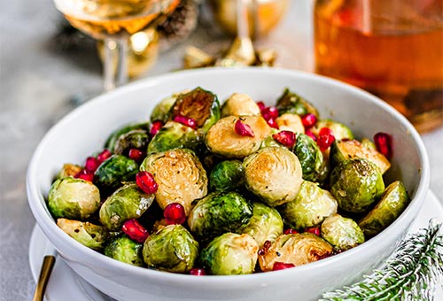 gebratener Rosenkohl mit Granatapfelkernen angerichtet in einer weißen Schale. Halb gefüllte Weingläser im Hintergrund.