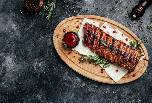 Spareribs liegen auf einer Holzplatte, daneben eine Soße mit Rosmarinzweige